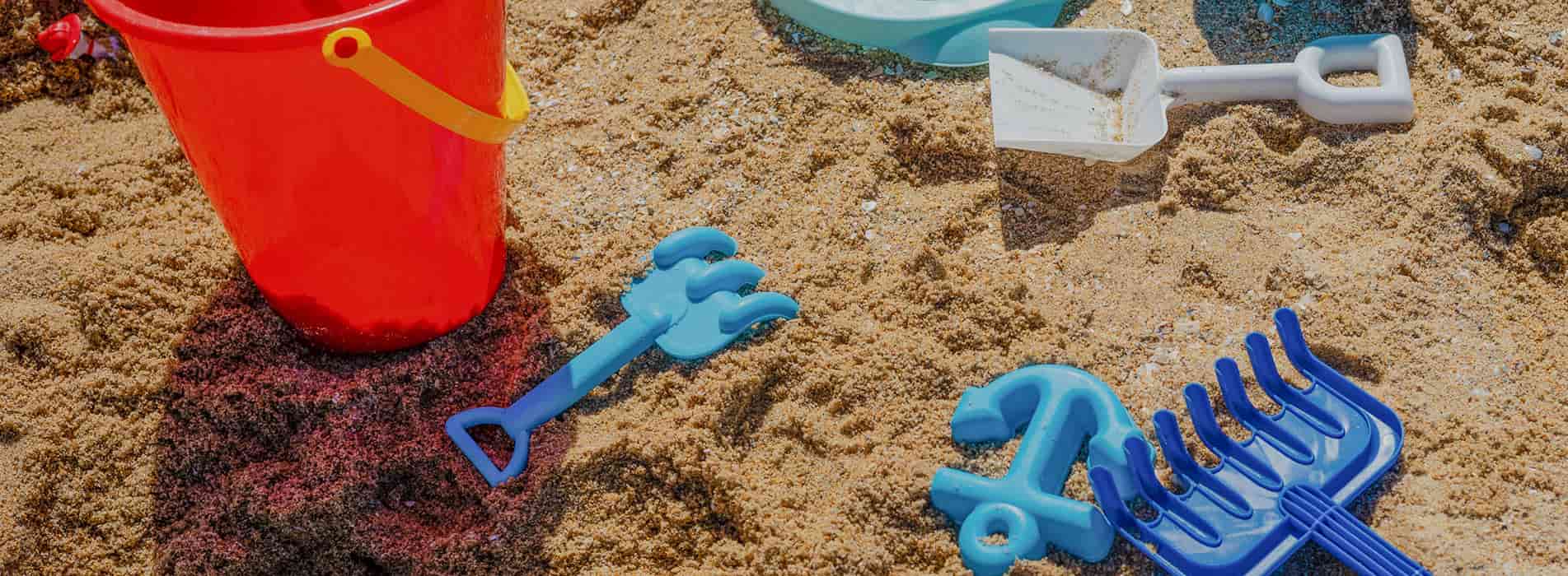 Colorful sand toys including a red bucket, blue shovel, and rake on sandy ground.